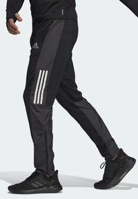 Svarta träningsbyxor med en slät yta, med grå sidopaneler, tre vita vertikala ränder och blixtlås vid vristerna. Bärs med svarta sneakers.