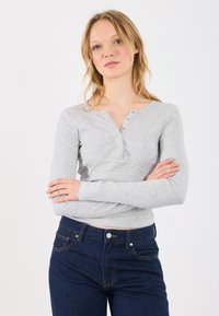 Svijetlosiva dugih rukava henley majica napravljena od rebrastog materijala s okruglim izrezom i tri dugmeta, uparen s tamnoplavim farmerkama.