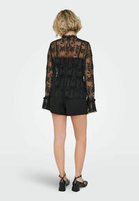 Černá krajková blůza s vysokým výstřihem, průsvitný materiál, květinová výšivka, dlouhé rukávy s volány, kombinována s černými šortkami.