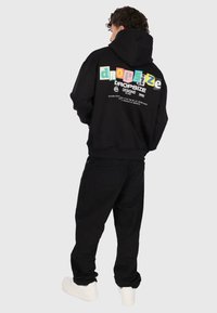Personne portant un sweat à capuche noir avec un logo « Dropsize » coloré dans le dos, un pantalon noir et des baskets blanches, se tenant devant un fond gris uni.