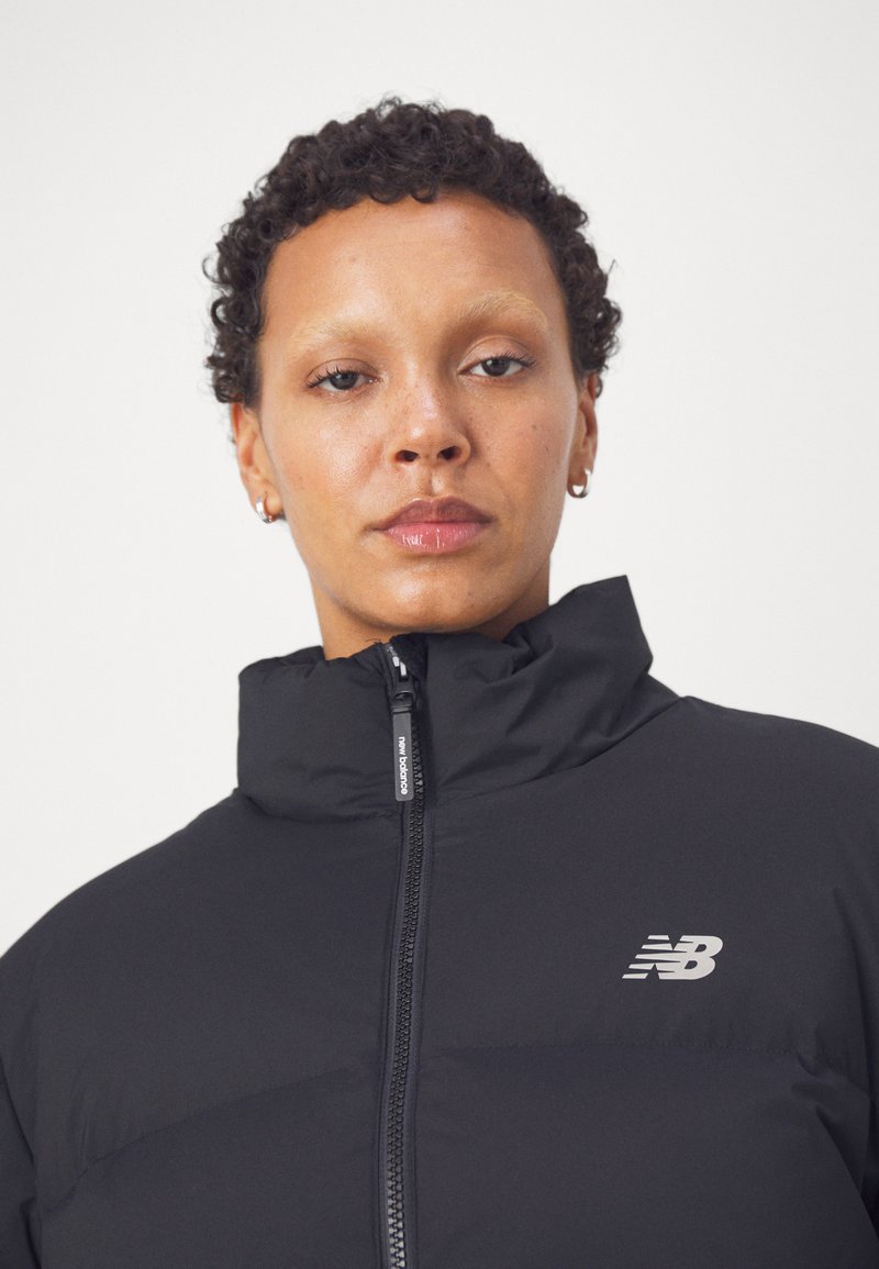 Person mit kurzen lockigen Haaren, die eine schwarze New Balance Daunenjacke bis zum Hals zugezippt trägt, vor einem schlichten weißen Hintergrund.