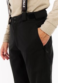 Close-up van een persoon in zwarte broek met ritszak en verstelbare tailleband, hand gedeeltelijk in de zak, beige met patronen lange mouw top.