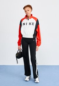 Biela, červená a čierna zipová bunda s logom "NIKE", skombinovaná s čiernymi nohavicami so bielymi bočnými prúžkami a šedými športovými topánkami. Malá čierna taška.
