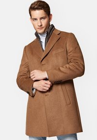 Brązowy płaszcz wełniany o strukturalnym kroju, z wyciętym kołnierzem, bocznymi kieszeniami i gładką fakturą. Noszony na szarej kurtce z zamkiem błyskawicznym.