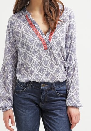 Donna che indossa una blusa bianca a maniche lunghe con motivi geometrici blu e dettagli rossi, abbinata a jeans blu scuro.