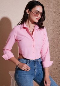 Pink cropped button-up shirt with long sleeves, featuring a pointed collar and visible buttons. Paired with blue high-waisted jeans.