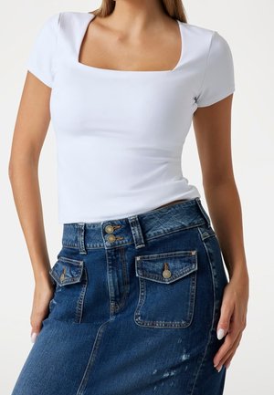 Mujer que lleva una camiseta blanca de cuello cuadrado y manga corta y una falda vaquera azul con bolsillos delanteros abotonados.