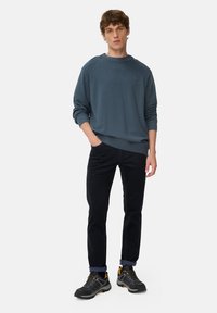 Bluoszary sweter z ściągaczami na rękawach, w połączeniu z czarnymi spodniami i szarymi trampkami z żółtymi akcentami. Model stoi z rękami w kieszeniach.