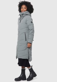 Manteau d'hiver gris long en doudoune avec un motif matelassé, capuche réglable, poches zippées à l'avant et zips sur les côtés, associé à une robe noire à côtes et des bottes épaisses.