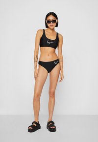 Set bikini nero con top a scollo rotondo e culotte coordinate con logo bianco. Il modello indossa occhiali da sole neri e sandali neri chunky.