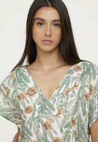 Blouse à imprimé floral avec un décolleté en V plongeant, des manches courtes, et un mélange de feuilles vertes et de fleurs jaune-orange sur fond blanc.