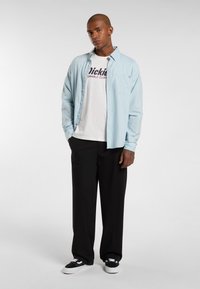 Svetle modrá košile na knoflíky přes bílé tričko s logem "Dickies", doplněná o černé široké kalhoty a černé tenisky s bílými podrážkami.