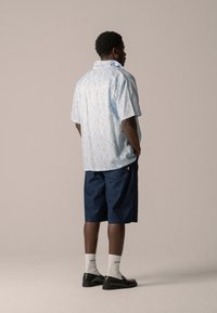 Lichtblauwe overhemdo met een wolkenpatroon, gecombineerd met donkere denim short. Model draagt zwarte schoenen en witte sokken.