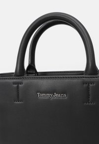 Svart läderhandväska med två rundade handtag, med en sydd design och en silverfärgad "Tommy Jeans"-logotyp på framsidan.