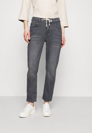 Femme portant un jean gris à cordon, des baskets blanches et un haut beige clair à manches longues, debout devant un fond neutre.