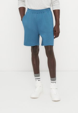 Egy férfi kék hétköznapi rövidnadrágot, fehér magas szárú tornacipőt, szürke-fehér csíkos zoknit és fehér hosszú ujjú inget visel, miközben egy fehér háttér előtt áll.