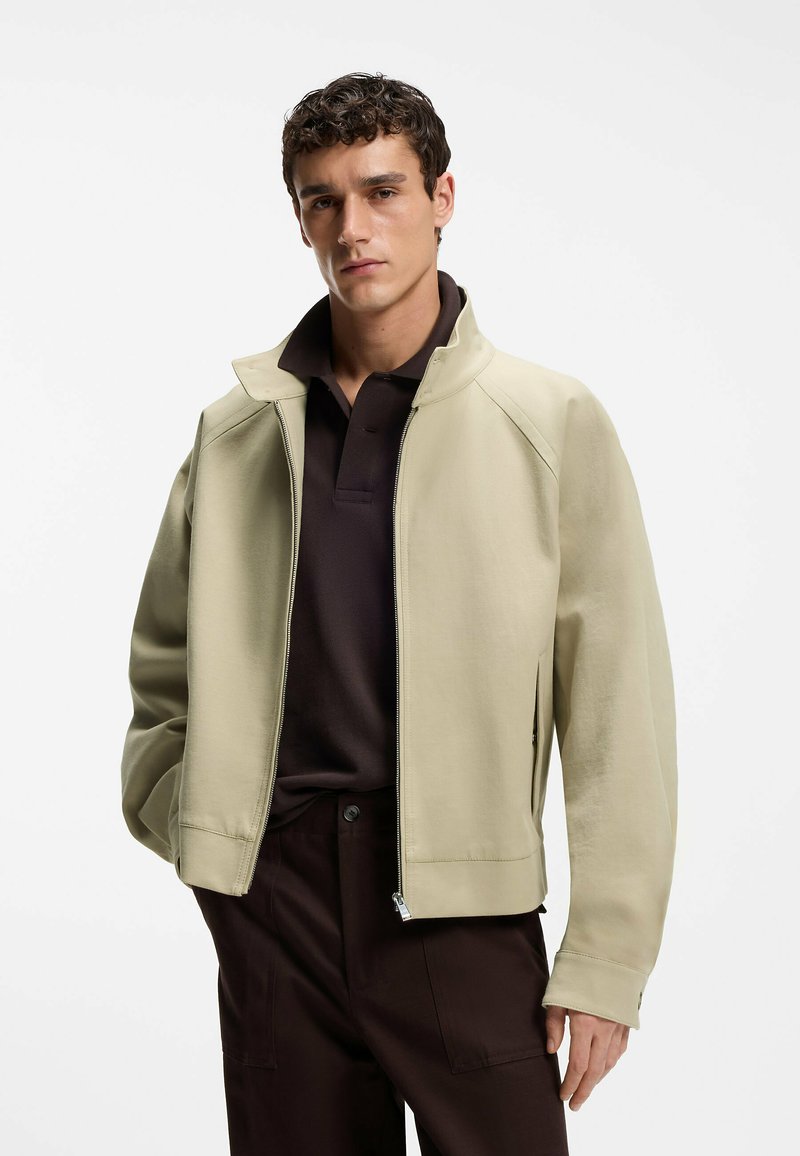 Jeune homme portant une veste beige zippée sur un polo noir et un pantalon marron foncé, debout devant un fond blanc uni.