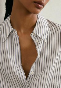 Valge ja meresinise triibuline nööbitav särk terava kraega, mis paljastab V-kaeluse. Sile kangas, tekstuuriga triibud, nähtavad nupud.