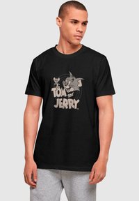 Mustvalge puuvillane T-särk, millel on vintage-stiilis halltoonides graafika Tomist ja Jerryst, allpool tegelaskujusid on julge tekst "Tom ja Jerry".