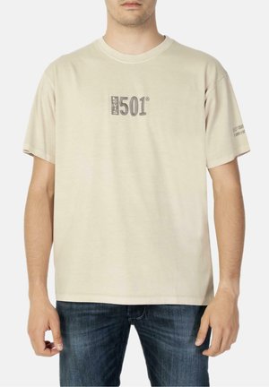 Beige Levi's 501 t-shirt båret af en mand, med "Levi's 501" logo på brystet og lille tekst på højre ærme, kombineret med blå jeans.