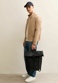 Giovane in giacca beige, jeans blu, sneakers bianche e berretto blu navy, che tiene un grande zaino nero a chiusura superiore per il manico.