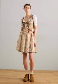 Robe dirndl beige à lacets avec un corsage ajusté, accents en dentelle, blouse blanche à manches courtes et un nœud à la taille, assortie à des chaussures à plateforme marron.