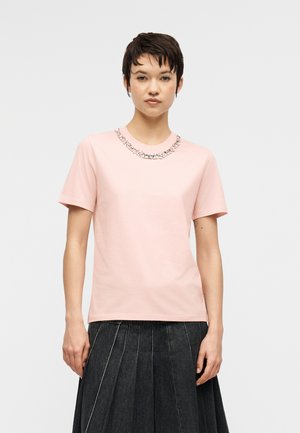 Donna con capelli corti e scuri che indossa una maglietta a maniche corte rosa chiaro con decorazioni argento sullo scollo e una gonna scura plissettata.