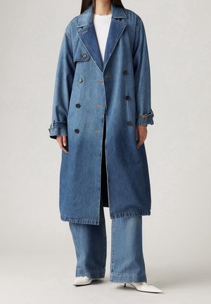 Personne portant un long manteau en denim à double boutonnage avec des détails de boutons, un jean à jambes larges, un top blanc et des talons blancs pointus.