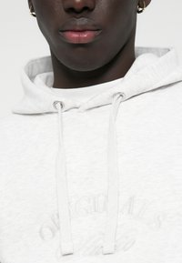Haut à capuche gris clair avec une texture douce, présentant une poche avant, des cordons de serrage et un logo en relief près de l'ourlet.