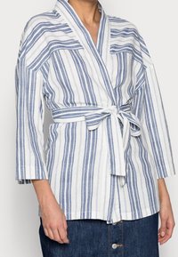 Raidallinen pellavapusero valkoisen ja sinisen sävyissä, jossa on kolmanneksen pituiset hihat, vyötäröllä sidottava vyö ja kaulus.