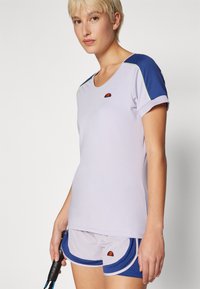 Camiseta deportiva de color púrpura claro con mangas azul marino y un pequeño logo. Combinada con pantalones cortos grises con acentos en azul marino y dobladillos redondeados.