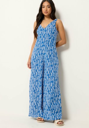 Kvinde modellerer ærmeløs blå jumpsuit med hvidt tov mønster, V-udskæring, vide ben og beige sandaler mod en ensartet lys baggrund.