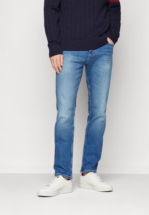 Personne portant un jean bleu, des baskets blanches et un pull bleu marine foncé, une main dans la poche, devant un fond uni.