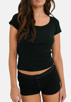 Młoda kobieta ubrana w czarną krótki rękaw z koronkowym wykończeniem oraz dopasowane czarne szorty z guzikami na tle jednolitego białego tła.