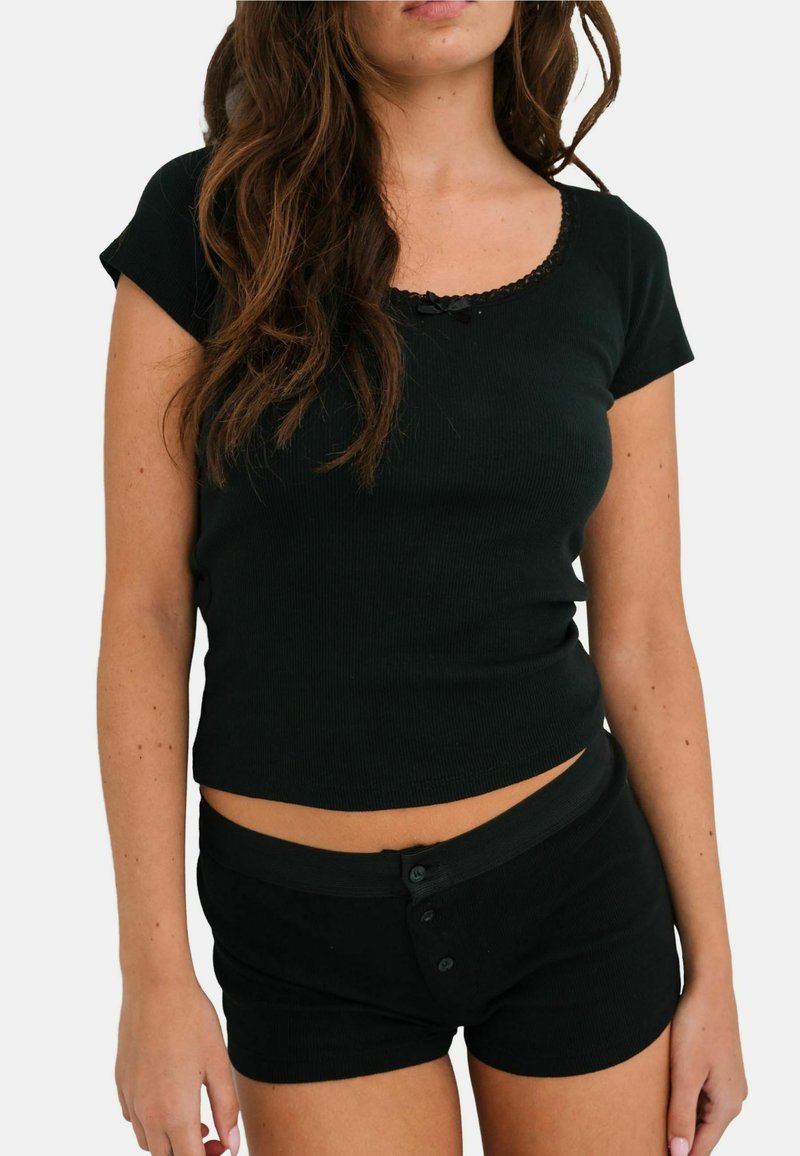 Jeune femme portant un haut noir à manches courtes avec bordure en dentelle et un short noir assorti à boutons, sur un fond blanc uni.