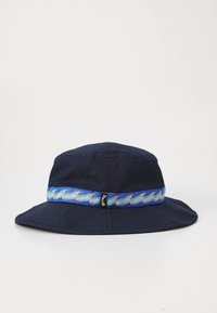 Tmavě modrý klobouk typu bucket s širokým krempou, zdobený barevným páskem s vlnkovým vzorem a malou logem na zadní straně. Bavlněný materiál.