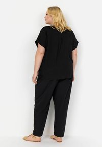 Haut noir ample à manches courtes, en tissu texturé, avec des plis dans le dos, associé à un pantalon noir et des sandales plates dorées.