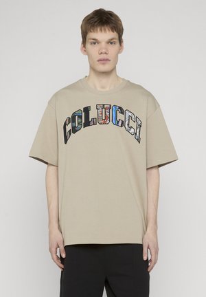 Young man wearing beige oversized T-shirt with "COLUCCI" text in colorful patterned letters, paired with black pants, standing against gray.