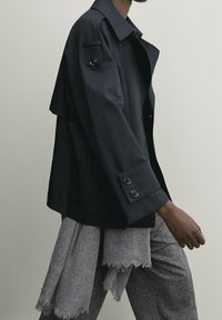 Granatowa kurtka o strukturalnym wzorze, z dużymi guzikami i boczną klapką. Noszona na jasnoszarym szaliku z postrzępionymi brzegami oraz dopasowanych spodniach.