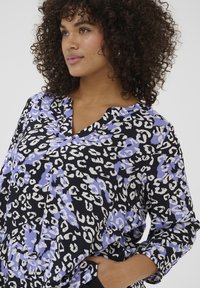 Schwarze Bluse mit einem Leo-Print-Muster in Lila und Weiß. Hat einen V-Ausschnitt und lange Ärmel mit gerafften Bündchen. Weicher Stoff.