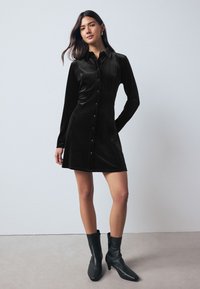 Černé sametové šaty košilového střihu s límečkem, dlouhými rukávy a předním zapínáním na knoflíky. Kombinované s černými kotníkovými botami, s přiléhavým živůtkem a rozšířenou sukní.