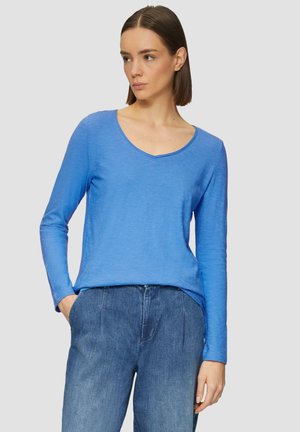 Femme avec des cheveux bruns raides portant un haut bleu à col en V à manches longues et un jean bleu ample, une main dans la poche, regardant à gauche.