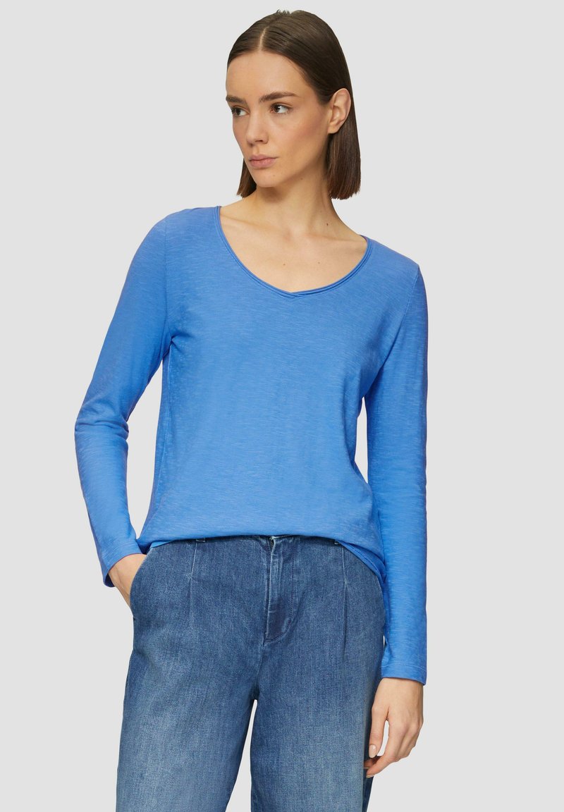Femme avec des cheveux bruns raides portant un haut bleu à col en V à manches longues et un jean bleu ample, une main dans la poche, regardant à gauche.