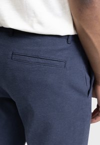 Pantalon texturé bleu marine avec une poche arrière à couture cachée. Le tissu a un motif subtil et une coupe ajustée.
