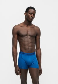 Άνδρας χωρίς πουκάμισο με μυώδη σωματότυπο, φορώντας μπλε boxer Jack & Jones, στέκεται μπροστά σε απλό ανοιχτό γκρι φόντο.