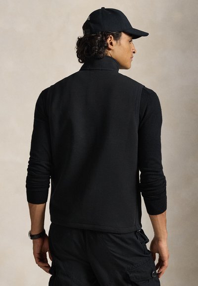 Μαύρο γιλέκο από φλις με ψηλό γιακά, μακριά μανίκια και ριμπ μανσέτες. Το υλικό είναι μαλακό, με λεία υφή. Φοριέται πάνω από σκουρόχρωμο παντελόνι καρό.