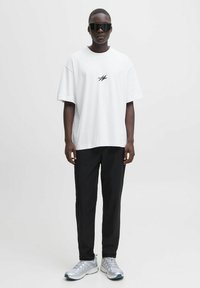 Mand iført oversize hvid t-shirt med sort logo, sorte bukser, sølvfarvede sneakers og store sorte solbriller, stående foran en ensfarvet baggrund.