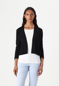 Zwarte cropped cardigan met sjaalkraag, lange mouwen en geribbelde structuur, laag over een witte top en gecombineerd met lichtblauwe broek.