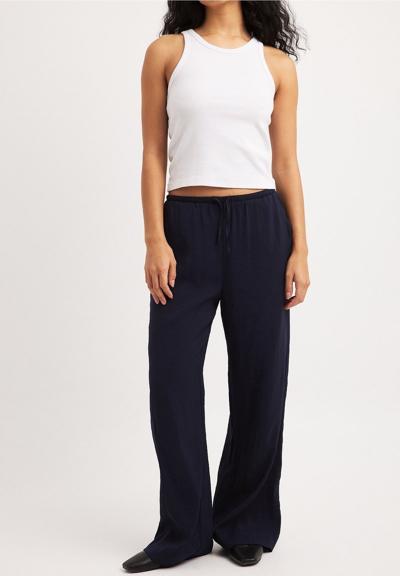 Femme portant un haut court blanc sans manches côtelé et un pantalon ample bleu marine avec cordon de serrage, ainsi que des chaussures noires, debout devant un fond uni.