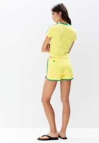 Geel kort aansprakelijk shirt met groene rand en logo, gecombineerd met bijpassende gele shorts met groene accenten en zijsplitten. Zwarte teenslippers.